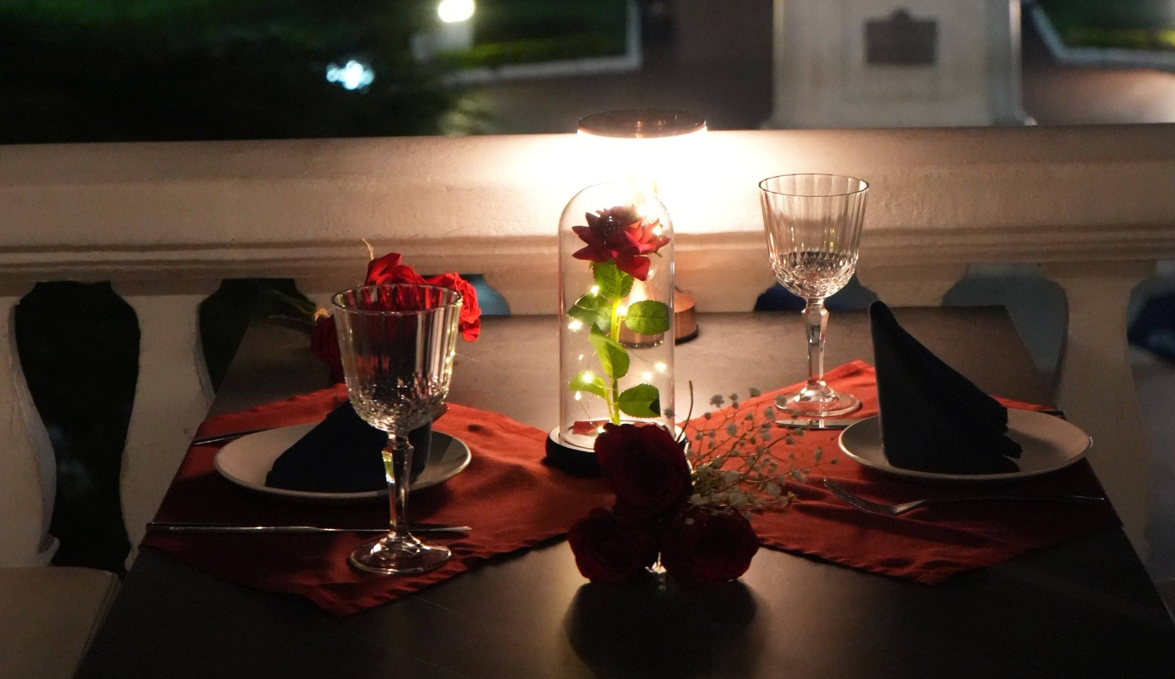 Mesa para dos en un balcón de noche, con una rosa roja bajo una campana de cristal, copas y servilletas negras